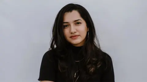 Kruthika Pillai Frizzle D'Souza wears a black blouse and sparkling necklace. The background is plain light gray.