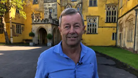 Norman Routledge smiles at the camera in front of the yellow painted entrance to the 18th century mansion at Ashton Court