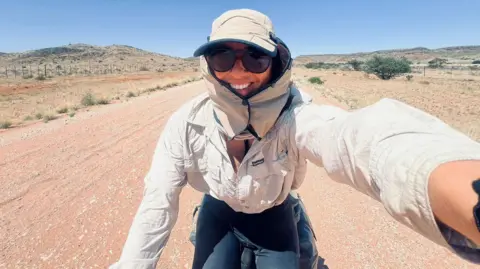 Ellie Mitchell-Heggs on her bike. Her head is covered with a hat with side panels and she is wearing a sand coloured shirt and black leggings. She is cycling on a sandy dirt road with dry greenery each side. There are hills in the background and she is smiling as she takes the selfie.