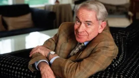 Stanley Baxter is photographed in later life.  He is looking at the camera wearing a suit and he has his arms crossed.