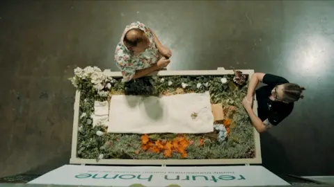 Ffilmiau Twm Twm Overhead shot, showing a human composting vessel filled with flowers and organic matter, with a small white sheet laid on the top. In front of the vessel is Kristopher Hughes, wearing a colourful shirt, and opposite at the end of the vessel is one of Return Home's staff members, a women dressed in a black t-shirt with her hair tied back