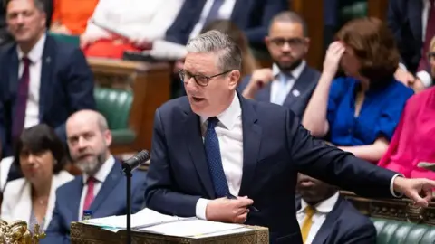 House of Commons Sir Keir Starmer in the Commons, pointing towards his colleagues on the frontbench. He is wearing a dark suit, white shirt and blue tie and leaning on the despatch box.