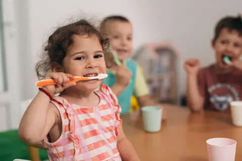 Getty Images Young children sat around a table brushing teeth. Image focuses on young girl with curly grown hair and striped peach tshirt, smiling as she brushes her teeth. There are pastel coloured plastic beakers on a wooden table and two young boys in the background who are also brushing their teeth