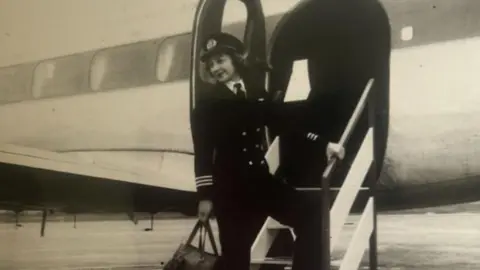 Candy Adkins A woman in a pilot outfit. She is standing on steps heading up towards a plane door. She is holding a bag. The image is black and white. 