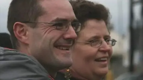 Other A smiling Peter Jenkins wears glasses and a grey coat. He is standing next to his smiling wife, Helen Greenham, who is also wearing glasses and has dark hair. 