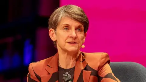 A head and shoulders shot of Laura McAllister. She has a handsfree headset on. There is a pink background behind her. She is wearing a tan and black suit and a black t-shirt underneath. She has a large pendant necklace on. She has short grey hair, with the fringe sweeping to one side. 