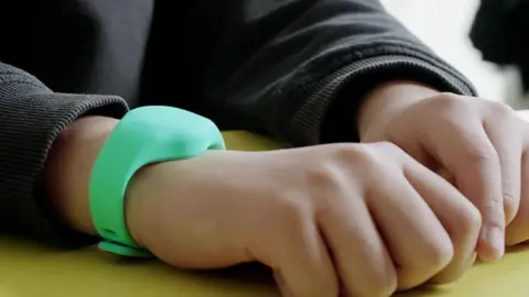 A small child wearing a black jumper has a mint-coloured wristband on his right wrist.