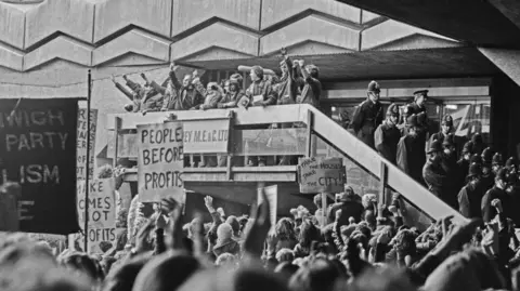 Getty Images A black and white picture of a protest. There are around 10 people standing on a balcony, some with their fists in the air. On the floor below, there are around 100 people looking up. Some are holding signs, with slogans reading: 'People before profits' and "Take the house, take the city'. There are also around 15 police officers standing on a staircase. 