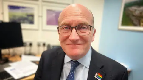Nikki Fox/BBC Nick Hulme with very short grey hair and glasses. He is smiling at the camera wearing a dark jacket, pale blue shirt and blue tie. There is a multicoloured flag emblem on his left shoulder. He is standing in an office with pictures on the wall and a large wooden desk on which a monitor is sitting.
