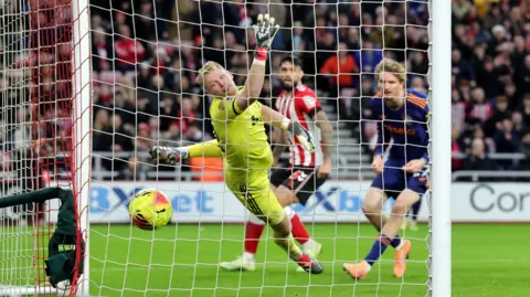Shot of behind a football goal with a yellow ball going into the net past a diving goalkeeper, wearing a yellow kit. The goalkeeper is looking at the ball. A player in an a blue top, Nick Woltemade, is crouched down looking at the ball. A Sunderland player is a red and white stripes is also in the background looking at the ball. In the background there are crowds of people all blurred out. 