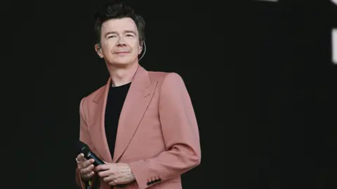 Rick Astley, wearing a pink blazer and black shirt, performing at Glastonbury Festival in July 2024