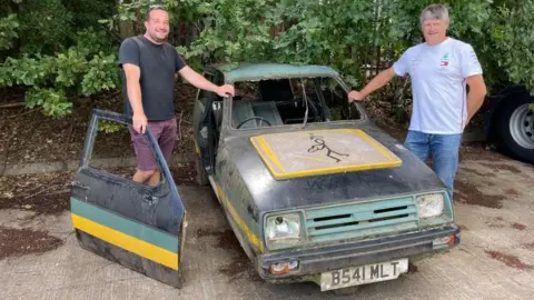 Dirty car painted in Saints colours - green roof and radiator grill, black bonnet with yellow and white rectangle in front of the window. The Saints stick man notif is on the rectangle. There is no glass in the windows.  The number plate B541 MLT is visible.  There are two men standing on either side of the car, with one hand on the chassis.  There are trees behind them.