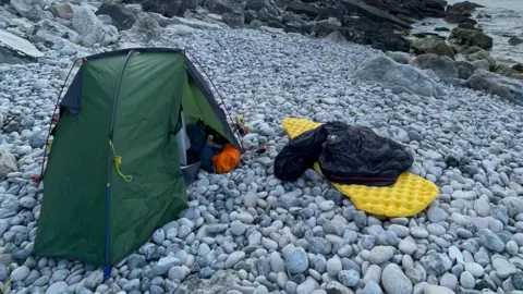 Mike Lambert A green tent next to a yellow roll-up camping bed mat laid out on grey pebbles on a beach