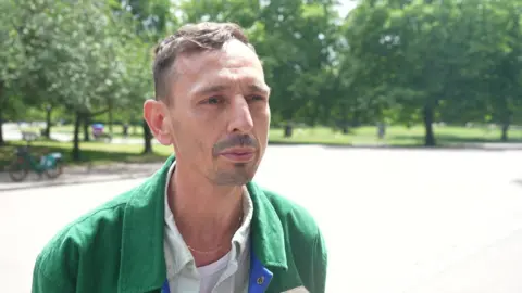 Gaz Kllokoqi, wearing a green jacket over a light-coloured shirt and a white T-shirt, speaks during an interview in Victoria Park, with trees and festival fencing in the background.
