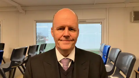 Ashley Fox is in the middle of the frame, wearing a grey suit and blue tie. He is looking at the camera with a neutral expression. Behind him are school chairs on a table. 