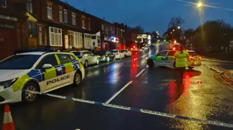 Phil Taylor Scene of crash - dark road with line of houses to left - police tape and orange cones cordon off the road. A police car is parked across the road. 