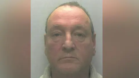 A police mugshot of a man in his sixties, with a reddish face and short grey hair. He is wearing a grey turtleneck jumper.