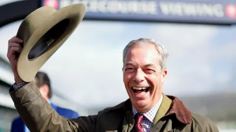 Getty Images Reform UK leader Nigel Farage in a wax jacket laughs as he lifts up his hat in front of a racecourse sign