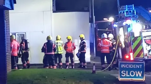 A group of firefighters standing between two houses. There is a lawn in front of them and to their right a fire engine. In front of that is a blue sign saying incident slow in white letters. It is night but the scene is well-lit.