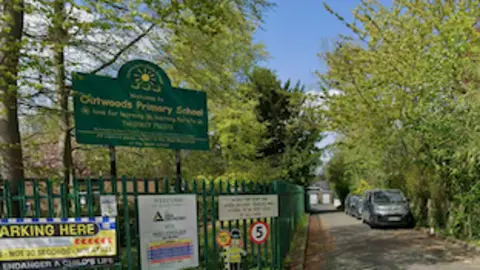 Entrance to Outwoods Primary School with green fencing, parked cars and trees lining the driveway.