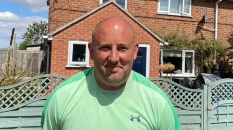 A man wearing a green t-shirt in front of a green fence and a brick house