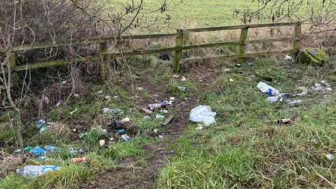 Image shows rubbish bottles and plastic debris in a lay-by. In the background behind a fence is a field with sheep in it.