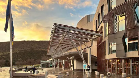 The front of the Holyrood building in deep red sunlight. The ground is wet and reflecting the light up and onto the light grey concrete facade.