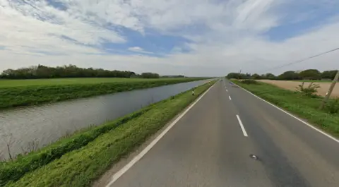 The road at Forty Foot bank in Ramsey, Cambridgeshire. It is a long straight road with a river running parallel alongside it. There are clouds above in the wide sky.