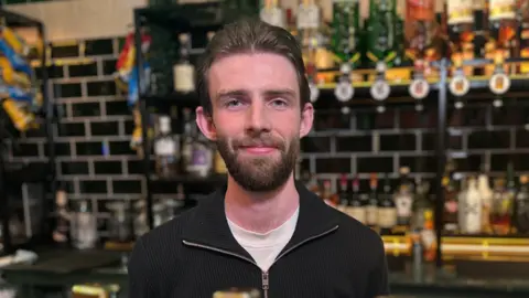 a smiling man with brown hair and wearing a black zipper jumper over a white t-shirt stands in front of optic bottles of spirits in a pub