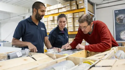 A man and a woman in navy tops stand next to a man in a red jumper as they look down at a wooden frame, where a rock desposit sits. In the background are stacked boxes in a warehouse