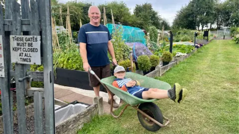 A man stands beside his allotment patch carrying a wheelbarrow. A young child is laid back in the wheelbarrow. They are both smiling.