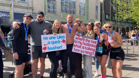 A group of friends, some of whom are wearing medals around their necks. Two are holding up signs - one reads "no one likes a quick finisher" another reads "where the hell is my husband what is taking him so long".