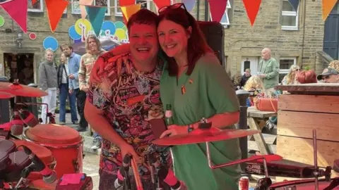 Hilary Bowyer (R) wearing a green dress and with her arm around Bongo Dave (L), who is wearing a patterned t-shirt at the event. They are stood next to musical instruments and people are stood looking at them behind them. 