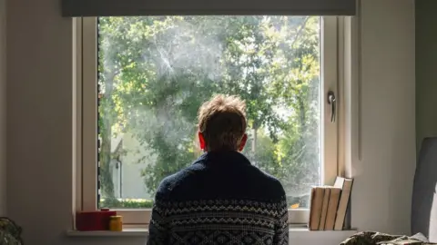 A stock image of a man with short dark hair wearing a blue patterned jumper looking out of a window. There are books on the window sill.