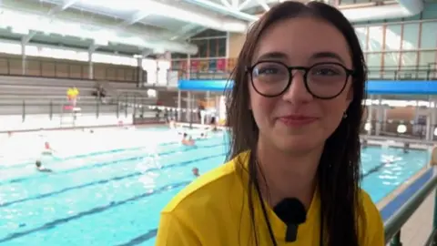 Blythe. A swimming pool can be seen behind her. She has long dark hair and glasses. She is wearing a yellow t-shirt. She is pictured smiling at the camera