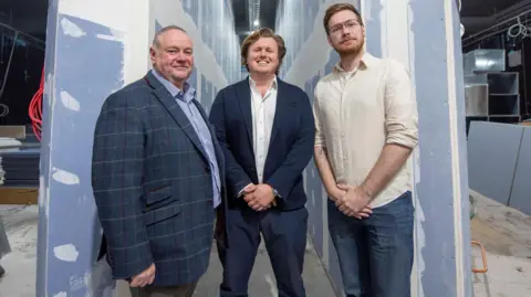 Three men stand in a line and smile at the camera. They stand in an area of the cinema being refurbished. The walls and blue with white paint marks on them and on the left a workman in a hi-vis jacket carries a plank of wood.