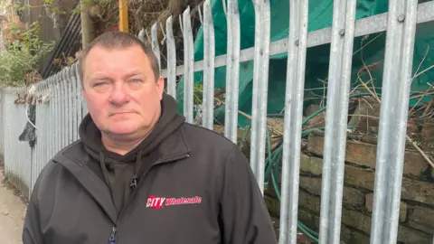 Malcolm Prior/BBC Geoff Howarth standing in front of iron railings, behind which sites the edge of the illegal waste site