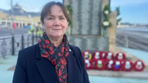 Diane Kelsey, who has brown hair and is wearing a navy coat with a poppy-themed scarf, standing in front of a war memorial on Douglas Promenade on the seafront that has poppy wreaths laid in front of it 