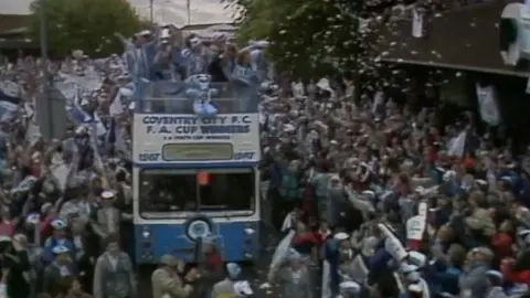 An open top blue bus with Coventry City FA Cup Winners on the front and people standing on top in flags and wearing hats. The bus is moving through a city street. Crowds around the bus raise their arms and throw confetti.