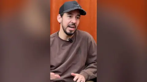 Mike Shinoda wearing a brown jumper and a black baseball cap. He's mid speech.