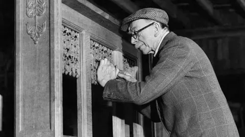 Getty Images A black and white photo of a man carving into wood. He wears a flat cap and a suit jacket and glasses, and has a moustache.