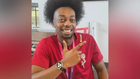 A man with Afro-style hair is wearing a red T-shirt with a rose motif and smiling at the camera. He is pointing to the sky with his right hand.