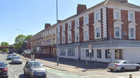 A large, smart-looking early 20th century building at a road junction. The ground floor is white and grey with signs advertising an engineering consultancy. The first and second floors are of red brick, with ornate white window frames. Cars pass on the roads outside.