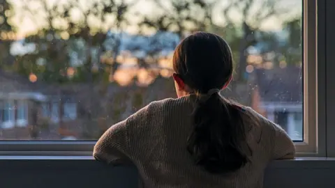 A young girl looks out of a window at a darkening sky. Seen from behind, she is wearing a jumper and has her hair in a ponytail