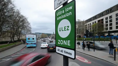 Green and white Ulez zone sign in foreground, cars in background