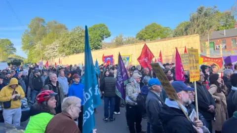 A crowded street, filled with people protesting against the Birmingham bin strikes. They are holding flags and placards. The sky is bright blue in the background.