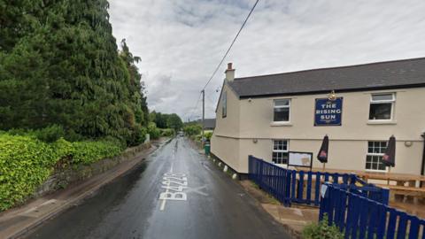 GV of the B4228 in Woodcroft. The road is close to a wall and trees to one side. The other side of the road is The Rising Sun Pub. It is a cream building with a blue fence.  