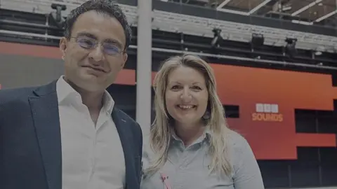 Surgeon Reza Arya stands next to Nicola Purdie in the BBC HQ in Cardiff. He has short, dark hair and wears glasses, a blue suit jacket and an open-necked white shirt. He is smiling. Nicola has long blonde hair. She is wearing a very light blue blouse. One large round drop earring can be seen in her left ear. They are in a studio with a red and orange panel behind them with the words "BBC Sounds" in BBC branding - three white squares containing the letters B,B,C in them in orange, and Sounds written underneath in light orange, all in capital letters.