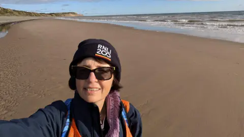 Anjie Rook Anjie Rook holding with her arm raised up taking a selfie on a sandy beach where the tide is out. She is wearing sunglasses and a navy blue hat with RNLI 200 on the front.