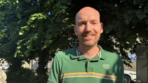 A smiling Christopher Pike in a green t-shirt with a grey and yellow band across the chest. He has a shaved head and a stubble beard.